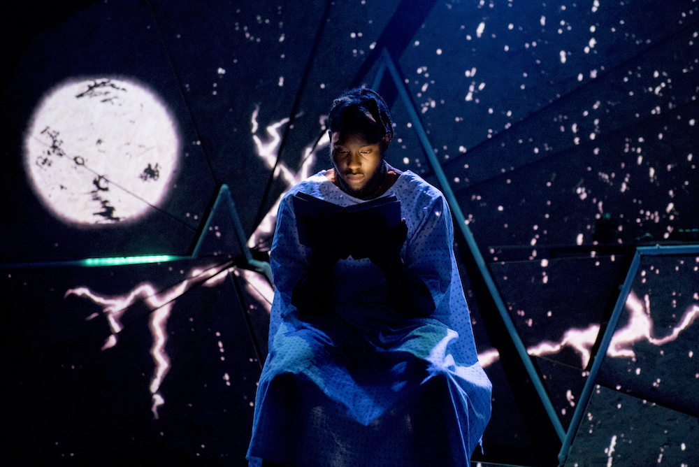 Caleb Femi pictured in front a full moon projected on a starry backdrop in Goldfish Bowl production
